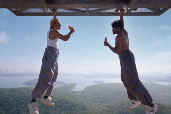 Two men are depicted hanging upside down from a metal structure against a clear blue sky. The man on the left wears a white tank top and a cap, holding a red bottle to his mouth. The man on the right is shirtless, also holding a red bottle. Both are wearing loose-fitting pants and sneakers, with a scenic backdrop of green forested hills and a body of water under a bright sky.
