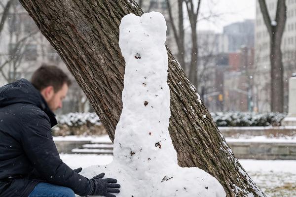 A man in a dark winter coat and gloves kneels next to a large, phallic snow sculpture built against the trunk of a tree. The ground is covered in snow, and the background features a blurry winter park setting.