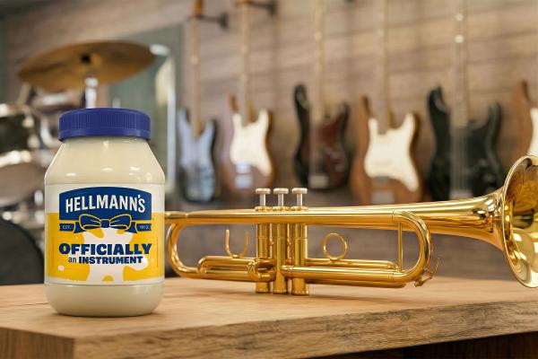 A Hellmann's mayonnaise jar with a blue lid and a label stating "Hellmann's OFFICIALLY an INSTRUMENT" is placed on a wooden table. Next to it is a gleaming golden trumpet. The background shows a collection of musical instruments, including guitars hanging on a wall and a drum kit, indicating a musical environment.