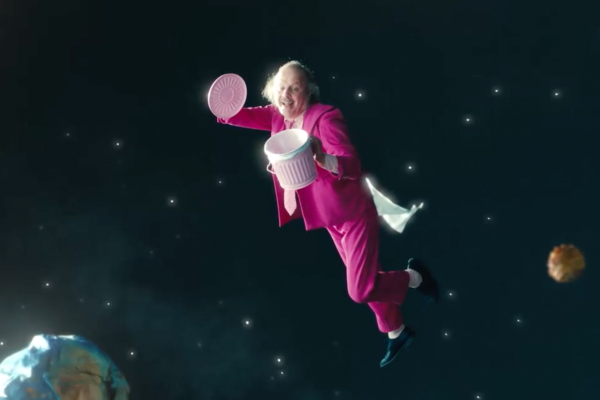 A cropped, standard-ratio version of French artist Philippe Katerine floating in outer space. Dressed in a vibrant pink suit and tie, he is captured mid-air, smiling as he holds open a small pink waste bin. A white wipe trails behind him, acting like a comet's tail amidst tiny, sparkling stars and a distant, stylized planet in the lower corner of the dark background.