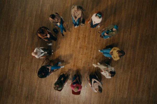 An overhead view shows a group of about a dozen people sitting in a circle on a wooden floor. They are all facing inward, and most appear to be sitting on chairs or stools. The floor is made of light brown wooden planks.