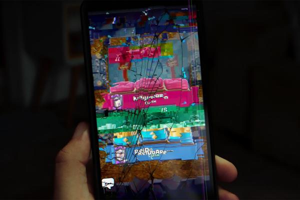 A hand holds a smartphone with a deeply cracked screen that also exhibits digital glitches and pixelation. The screen shows a distorted view of a mobile game, possibly "Clash Royale," with blocky, colorful graphics of game structures and characters visible through the extensive screen damage.
