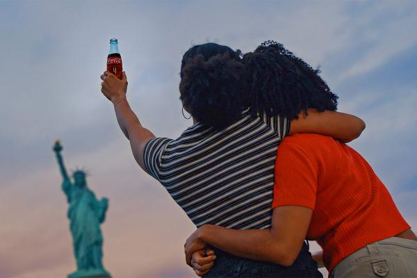 Two women with curly hair, seen from behind, stand arm-in-arm. One raises a glass Coca-Cola bottle towards the distant, out-of-focus Statue of Liberty against a cloudy sky.
