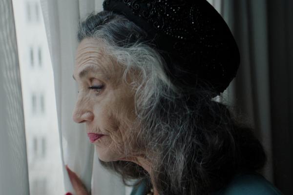A side profile shot of an older woman with grey and dark hair, wearing a black beaded hat and a teal top, gazing out a window. Her hand, with red fingernails, is visible holding a white curtain. Blurry city buildings are seen through the window in the bright light.