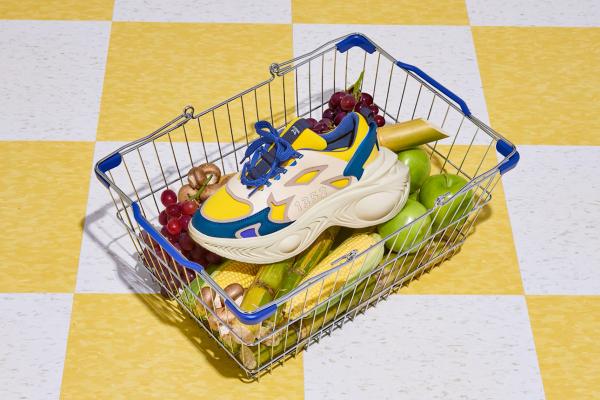 A grocery shopping basket sitting on a yellow and white tiled floor. The basket holds various fruits and vegetables, and on top lies a blue white and yellow sneaker