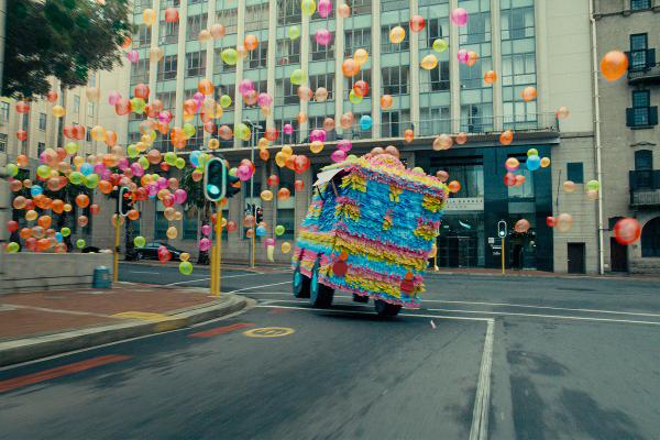 A car turrns down a street, in the background there are hundreds of colorful balloons
