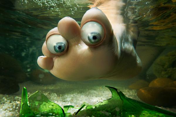 Underwater shot of a foot with eyeball-like toes near broken green glass and pebbles,