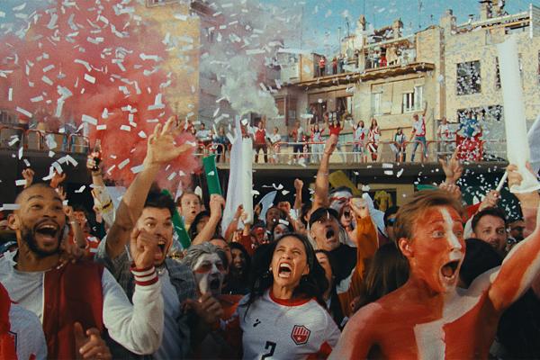 A large, passionate crowd of soccer fans celebrates enthusiastically outdoors in front of residential buildings. Many fans are shouting with joy, wearing red and white team colors, and some have red and white paint on their faces. White confetti falls from the sky, a red smoke flare is set off in the background, and inflatable noisemakers are held in the air.