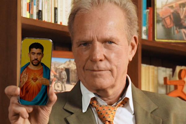 Close-up of an older man in a brown suit and orange tie holding a phone toward the camera. The screen shows a religious icon-style illustration of Bad Bunny with a sacred heart and halo.