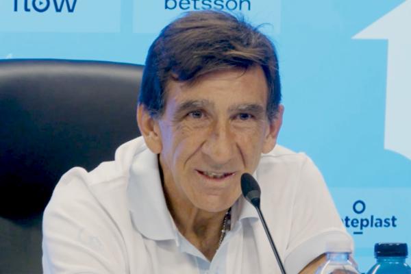 "A man in a white polo shirt speaks at a press conference microphone, with sponsor logos including Betsson and Flow visible on the blue backdrop behind him."