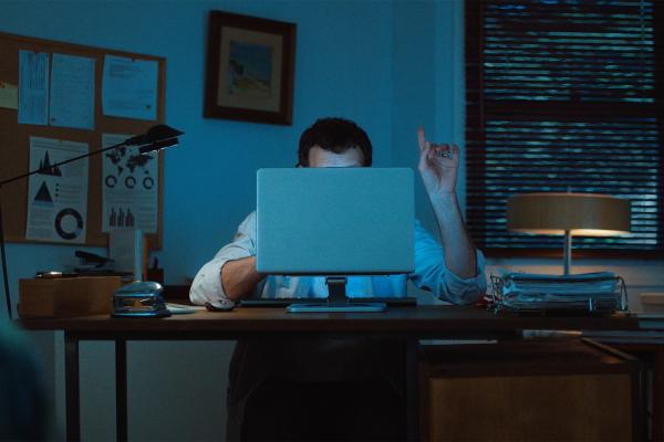 A man sitting at an office desk at night in dim blue lighting, hiding his face behind an open laptop monitor while raising his index finger in a "just one minute" gesture.