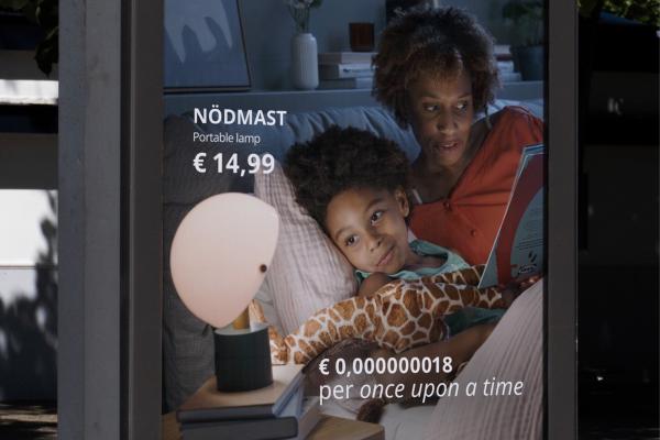 Close-up of an IKEA poster advertisement for the NÖDMAST portable lamp (€14.99). The photo shows a mother reading a bedtime story to her child in bed next to the illuminated lamp, with tagline text reading "€ 0,000000018 per once upon a time."