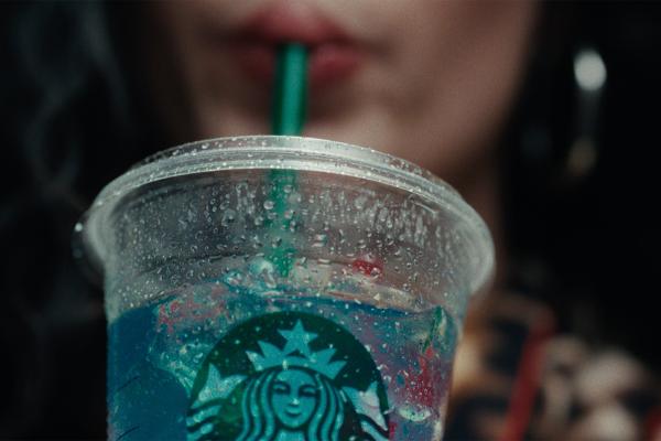 Macro shot of a cold, clear plastic Starbucks cup beaded with water droplets, featuring the iconic green Siren logo and a vibrant blue iced drink, with someone sipping through a green straw in the soft-focus background