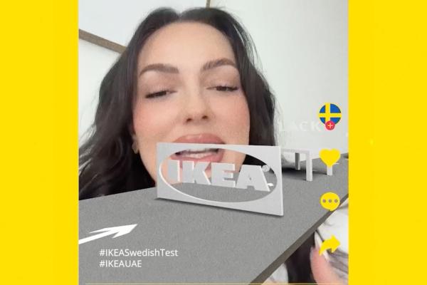 Mobile screen capture of a woman testing the interactive #IKEASwedishTest AR filter on TikTok, showing a 3D IKEA logo cutout and a miniature Lack side table gliding along a grey path accompanied by the Swedish flag icon and social UI buttons.