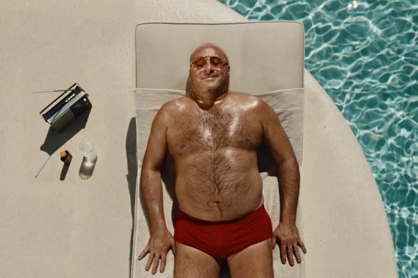 Direct overhead shot of a smiling, tanned bald man relaxing on a sunbed beside shimmering blue pool water, wearing red swim briefs, a gold chain, and tinted sunglasses, next to a vintage radio and a bottle of tanning oil.