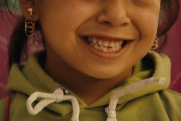 Cropped close-up focusing on the joyful grin of a young child wearing dangling gold hoop earrings and a sage-green hooded sweatshirt with white drawstrings.