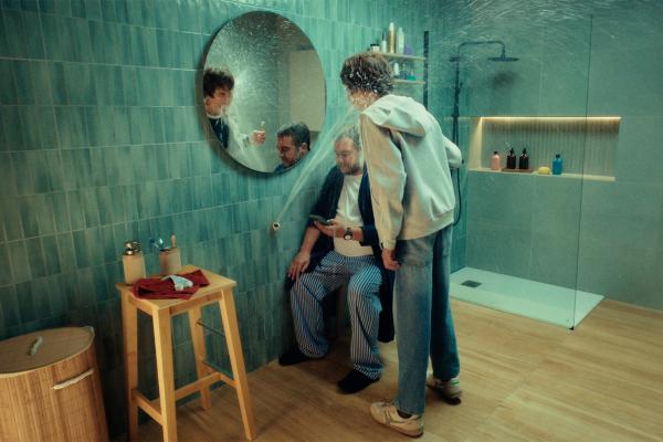 A humorous bathroom scene where a high-pressure jet of water shoots from an unfinished wall pipe against a round mirror while a boy with a toothbrush and his father in pajamas wait underneath.