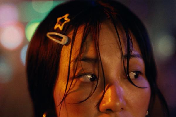Moody portrait of a woman looking sideways with rain-dampened hair and star hair clips, reflecting warm neon bokeh lights.
