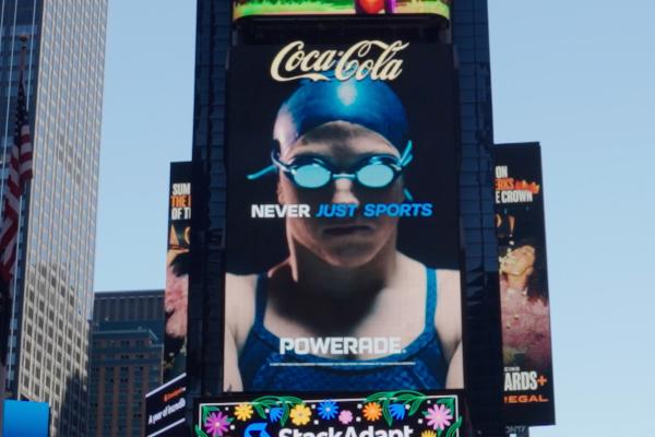 A Times Square electronic billboard featuring a close-up of a swimmer in mirrored goggles and swim cap with the text 'Coca-Cola', 'NEVER JUST SPORTS', and 'POWERADE'.
