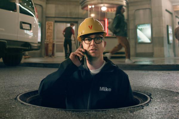 Man in a yellow safety helmet on a phone while emerging out of a manhole