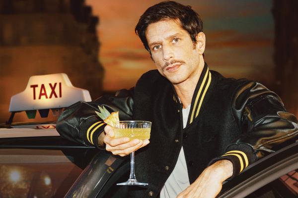 Young man holding a cocktail in one hand while leaning outside of the door of a taxi