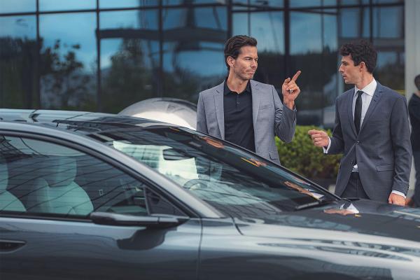 Two young men standing behind an Audi car. They are both wearing suits. The man on the left is point his finger at the man on the right.