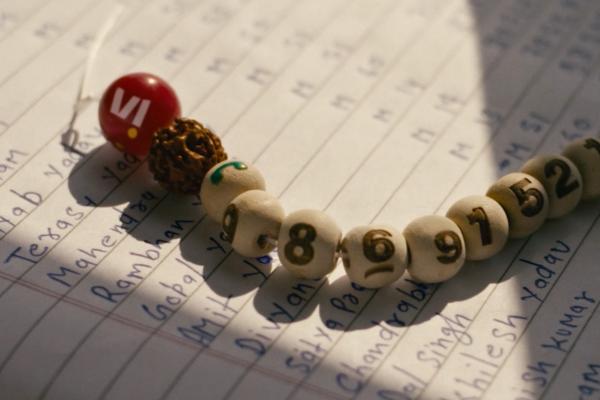 Close-up of a bracelet made of beads with numbers, including "8," "6," "9," "7," "5," and "2," resting on a lined paper with handwritten names. A red bead with the "VI" logo is also visible.