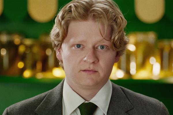 A fair-skinned man with blond, wavy hair and blue eyes looks directly at the camera with a neutral expression. He is wearing a grey suit, white shirt, and green tie, standing in front of a green and gold background.