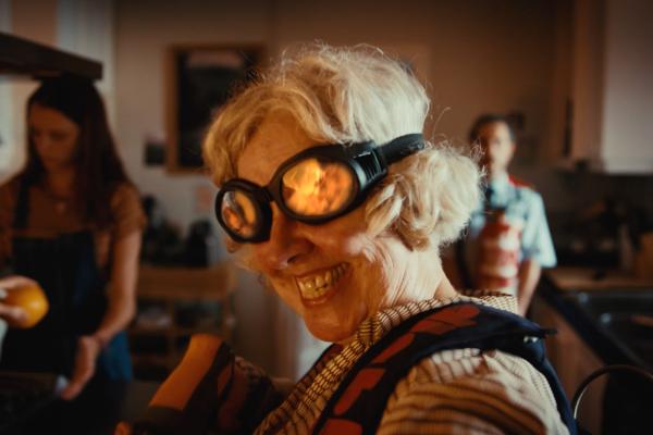 A close-up shot shows an older woman with blonde hair, wearing black goggles that reflect a fiery orange glow, smiling widely with her teeth showing. She is wearing a patterned shirt with a dark blue and red design. In the blurred background, another woman in a dark apron holds a yellow fruit, and a man in a light blue shirt and red apron stands further back.