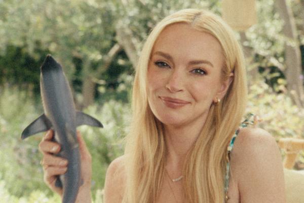 A smiling Lindsay Lohan, with long blonde hair, holds up a small, dark grey shark toy. She wears a patterned top and a delicate necklace. The background is blurred with green foliage.