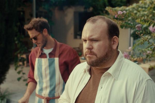 A close-up shot of two men in a garden setting. In the foreground, a man with a beard and a light-colored shirt looks directly forward with a serious expression. Behind him and slightly out of focus, another man wearing sunglasses and a striped apron is visible, looking down towards something out of frame.