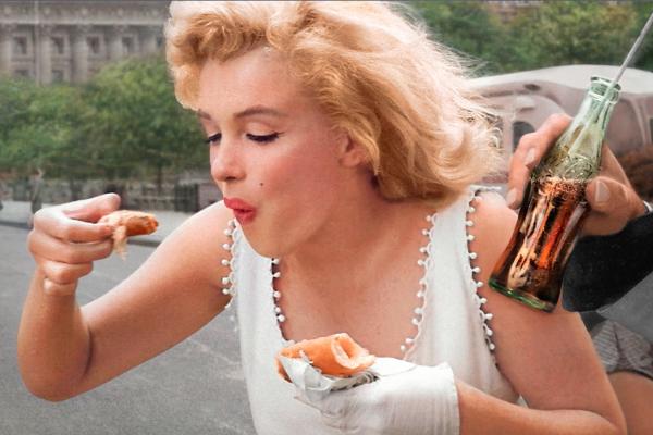 Marilyn Monroe eating while a hand holds a Coca-Cola bottle frame right