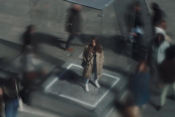 An overhead look of a woman standing alone in a glass cube while others are surrounding her from the outside