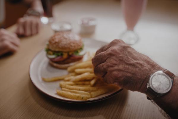 The arm of an old man picks at a plate of french fries and a burger on a plate at a restaurant