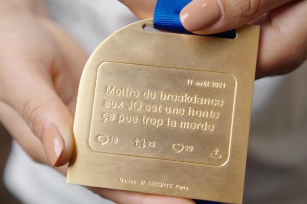Hands holding a gold medal with an engraving that says "Mettre du breakdance aux JO est une honte ça pue trop la merde" with social media engagement icons