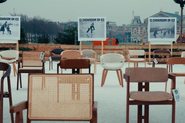 An outdoor ice rink is transformed into a showroom, featuring numerous modern chairs of different styles and colors arranged on the ice. In the background, large billboards advertise "SHOWROOM ON ICE" with images of people ice skating with chairs, and buildings are visible beyond the rink perimeter.