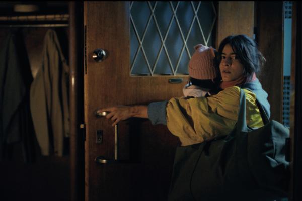 A woman with dark hair, dressed in a yellow jacket and red scarf, reaches to open a wooden front door with diamond-patterned glass. She is carrying a small child, whose head, wearing a pink beanie, is visible over her shoulder. An open wooden closet with clothes hanging inside is visible to the left, and the scene is dimly lit.