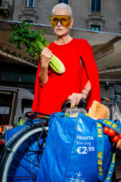Full portrait campaign visual featuring an elderly woman in an avant-garde red cape and yellow sunglasses holding celery, standing beside her bicycle where a blue IKEA FRAKTA bag is repurposed as a bike pannier, complete with the headline 'FRAKTA Bike bag €2,95'.
