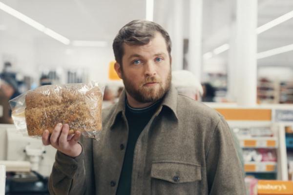 A man stands at a Sainsbury's supermarket checkout holding a loaf of bread