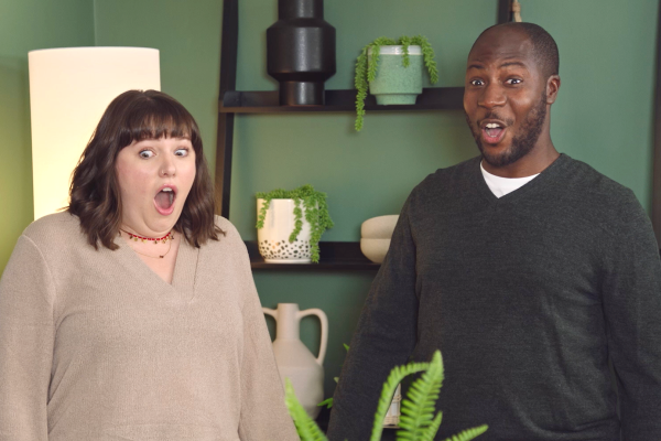 A woman and man stand in a green living room with surprised expressions on their faces