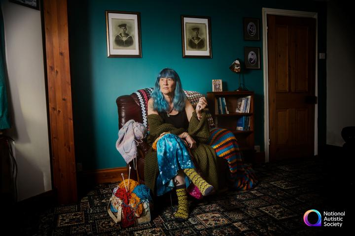 Woman with turquoise hair sits in arm chair in front of turquoise wall. She is surrounded by photographs and knitting materials.