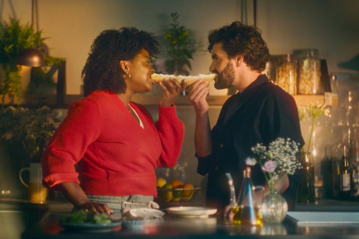 A man and woman bite into opposite sides of a baguette.